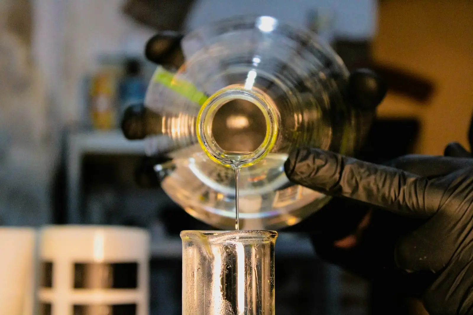 Mano con guante vertiendo un líquido en un vaso de precipitados, en un laboratorio de química con poca luz