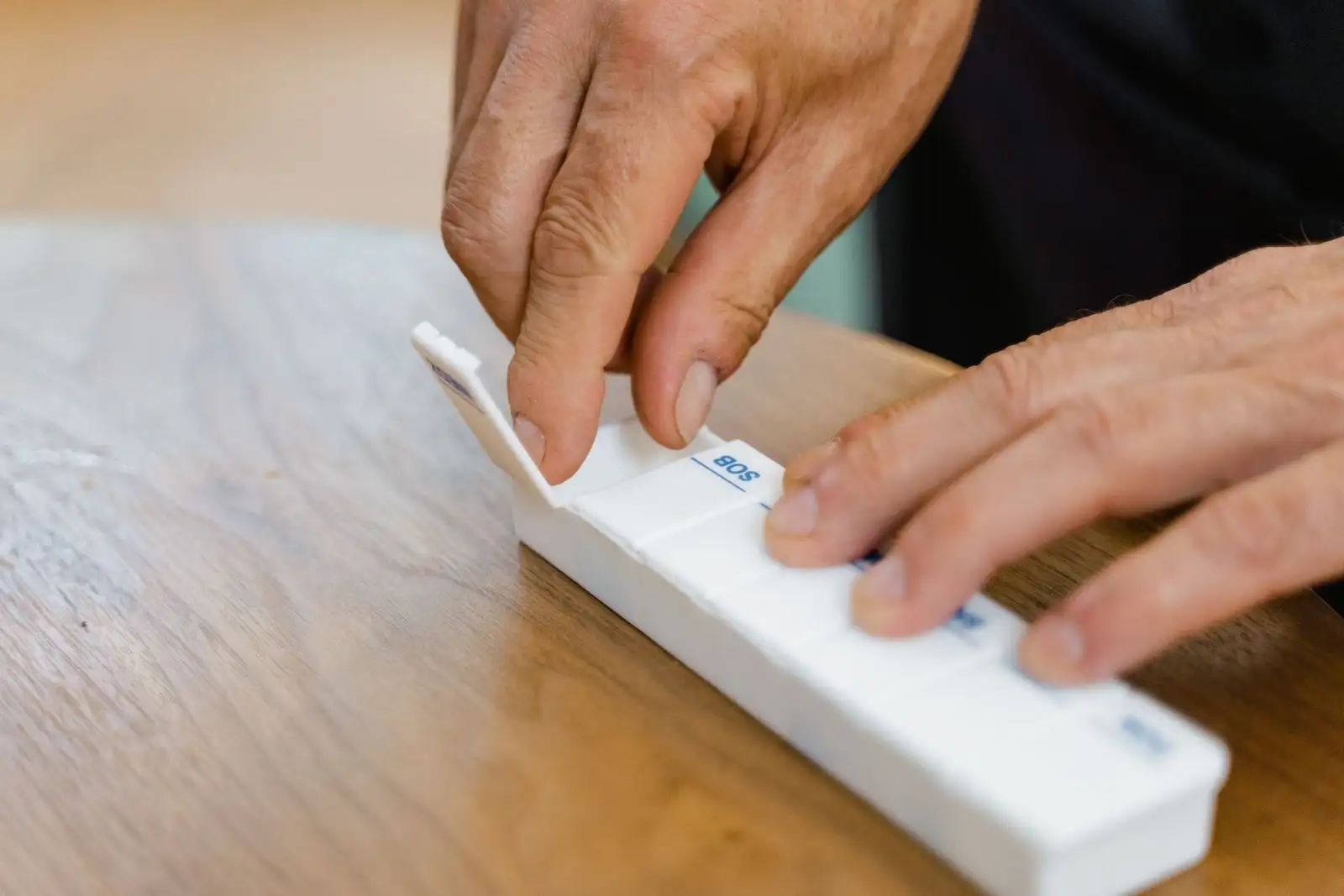 Manos de una persona mayor que reparte sus medicamentos en un pastillero semanal sobre una mesa de madera