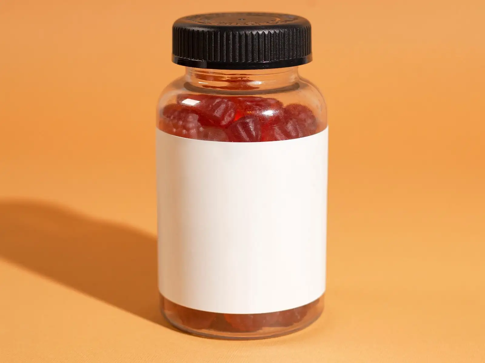 Bote transparente lleno de gominolas rojas, etiqueta en blanco, sobre fondo naranja
