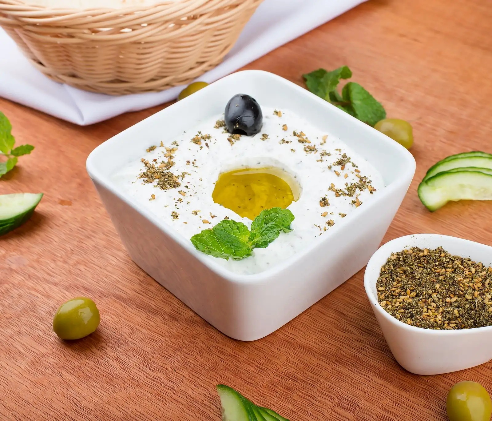 Tzatziki en un bol cuadrado blanco, con un chorro de aceite de oliva y menta, rodeado de rodajas de pepino y aceitunas