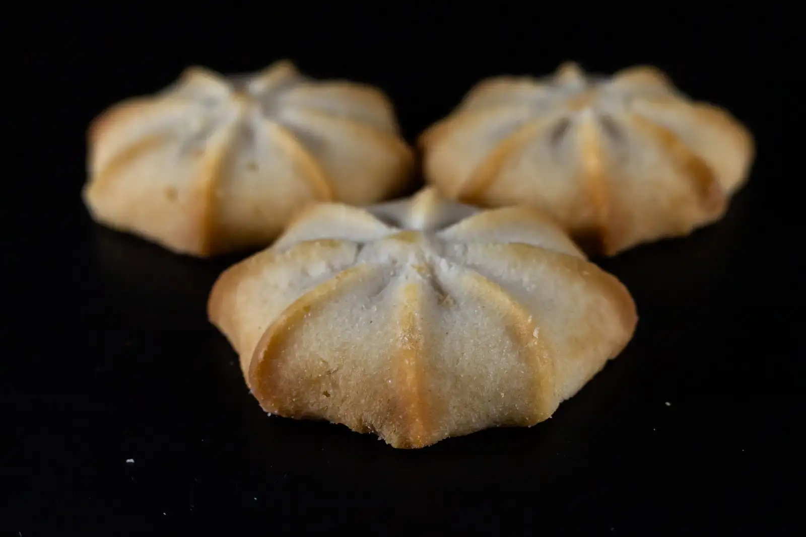 Tres galletas de mantequilla doradas en forma de roseta sobre un fondo negro