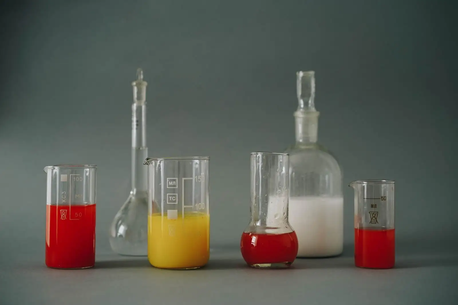 Vasos de precipitados y matraces de laboratorio con líquidos rojos, amarillos y blancos sobre fondo gris