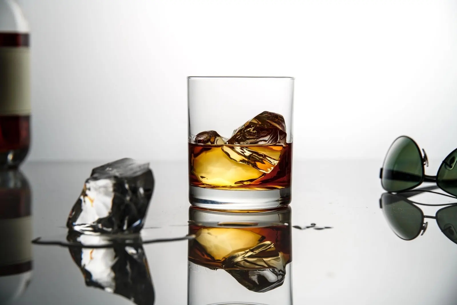 Vaso de whisky con hielo sobre una superficie reflectante, fondo blanco