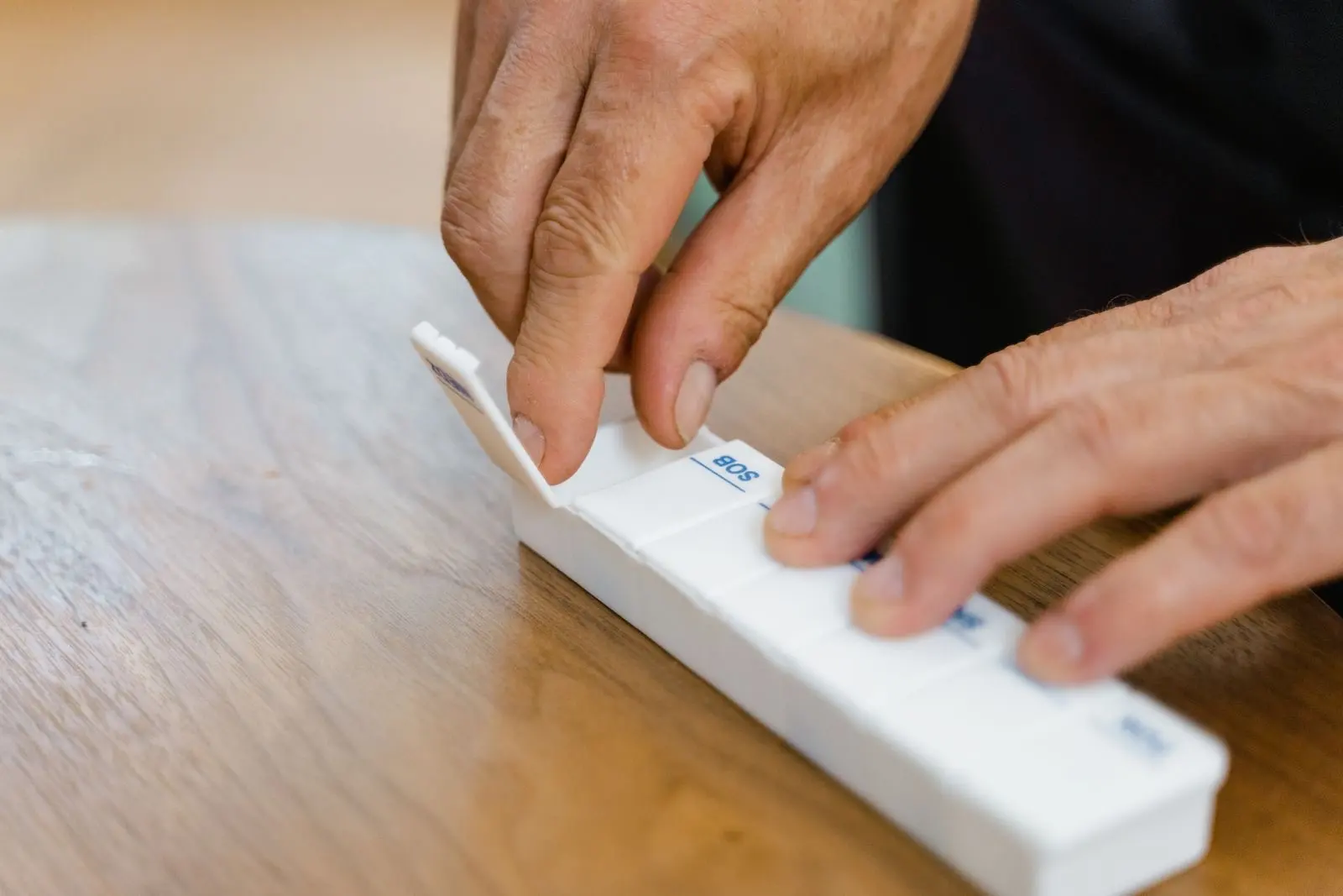 Manos de una persona mayor que reparte sus medicamentos en un pastillero semanal sobre una mesa de madera