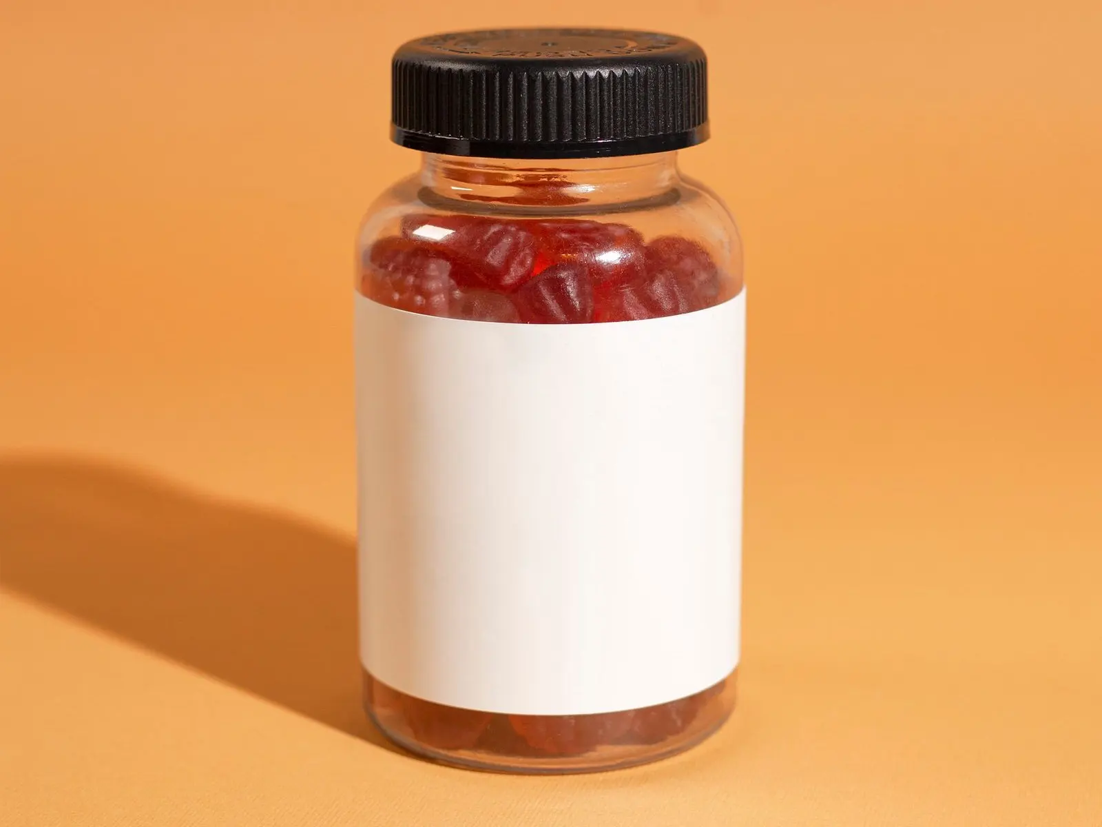 Bote transparente lleno de gominolas rojas, etiqueta en blanco, sobre fondo naranja
