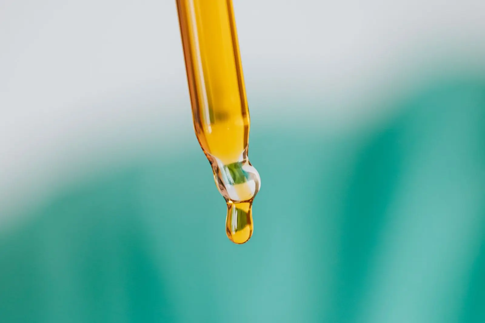 Pipeta de vidrio que deja caer una gota de aceite ámbar, sobre fondo verde desenfocado