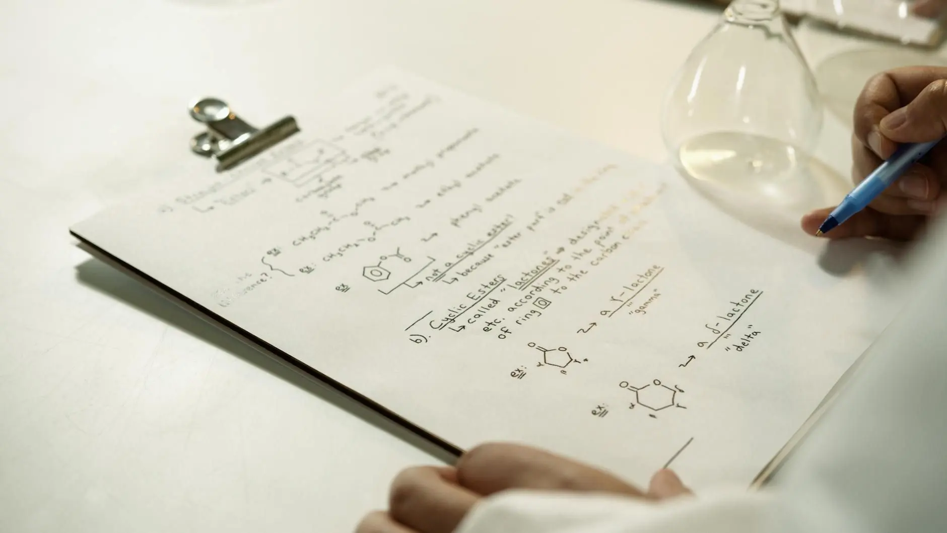 Apuntes de química orgánica escritos a mano en una carpeta con pinza, junto a material de vidrio de laboratorio